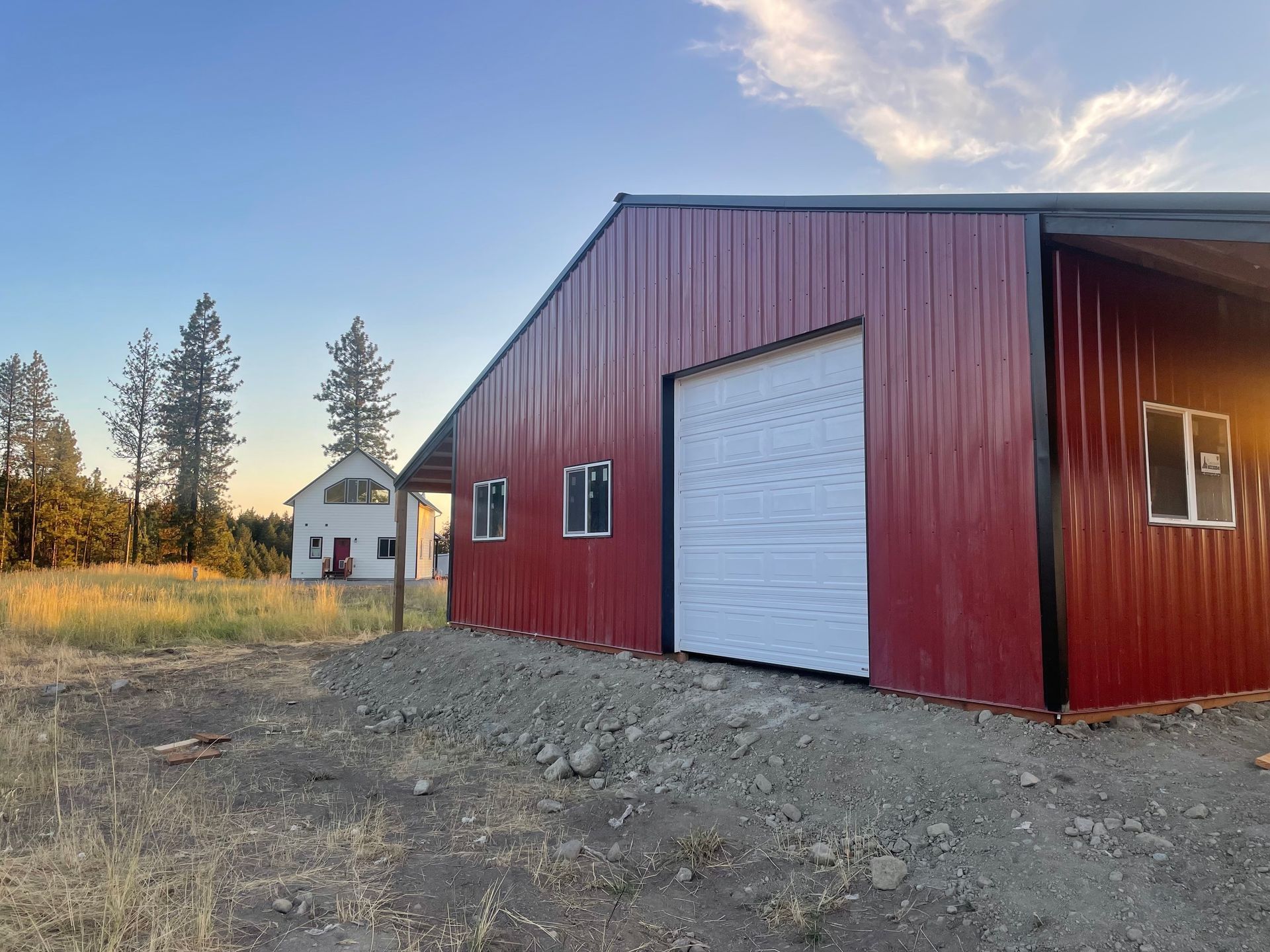 A red metal barn with a white garage door and two windows stands on a gravel lot next to a white house at sunset.