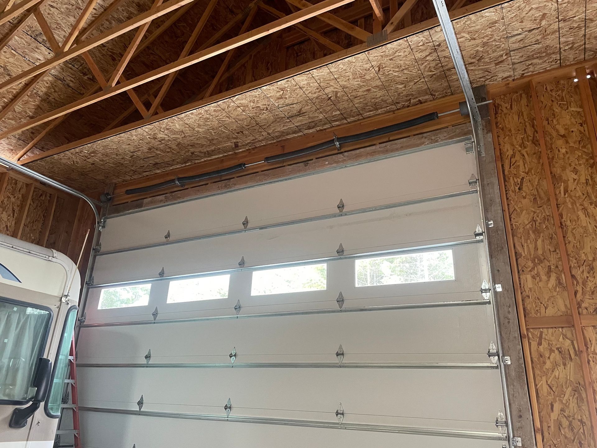 An indoor view of a white, sectional garage door with a row of four rectangular windows, installed in a wooden workshop.