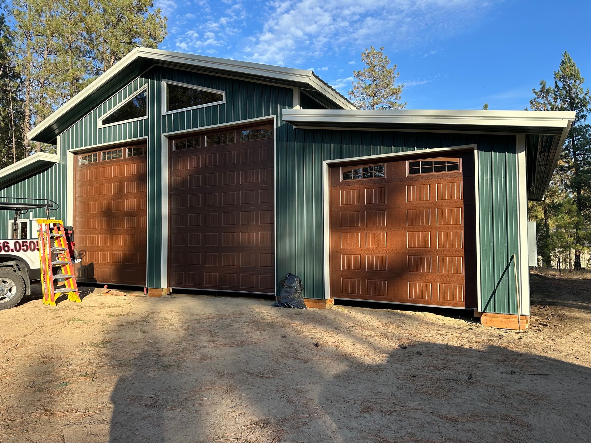 A green metal garage building with two large brown carriage-style garage doors in a wooded, dirt-covered outdoor setting.