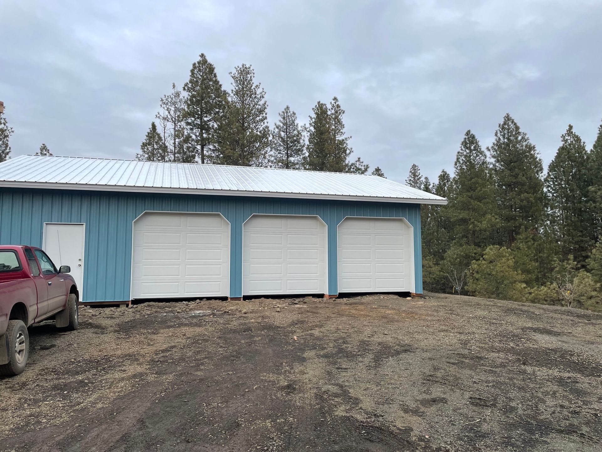 A blue detached three-car garage with a white roof sits on a gravel lot next to a red pickup truck, backed by pine trees.