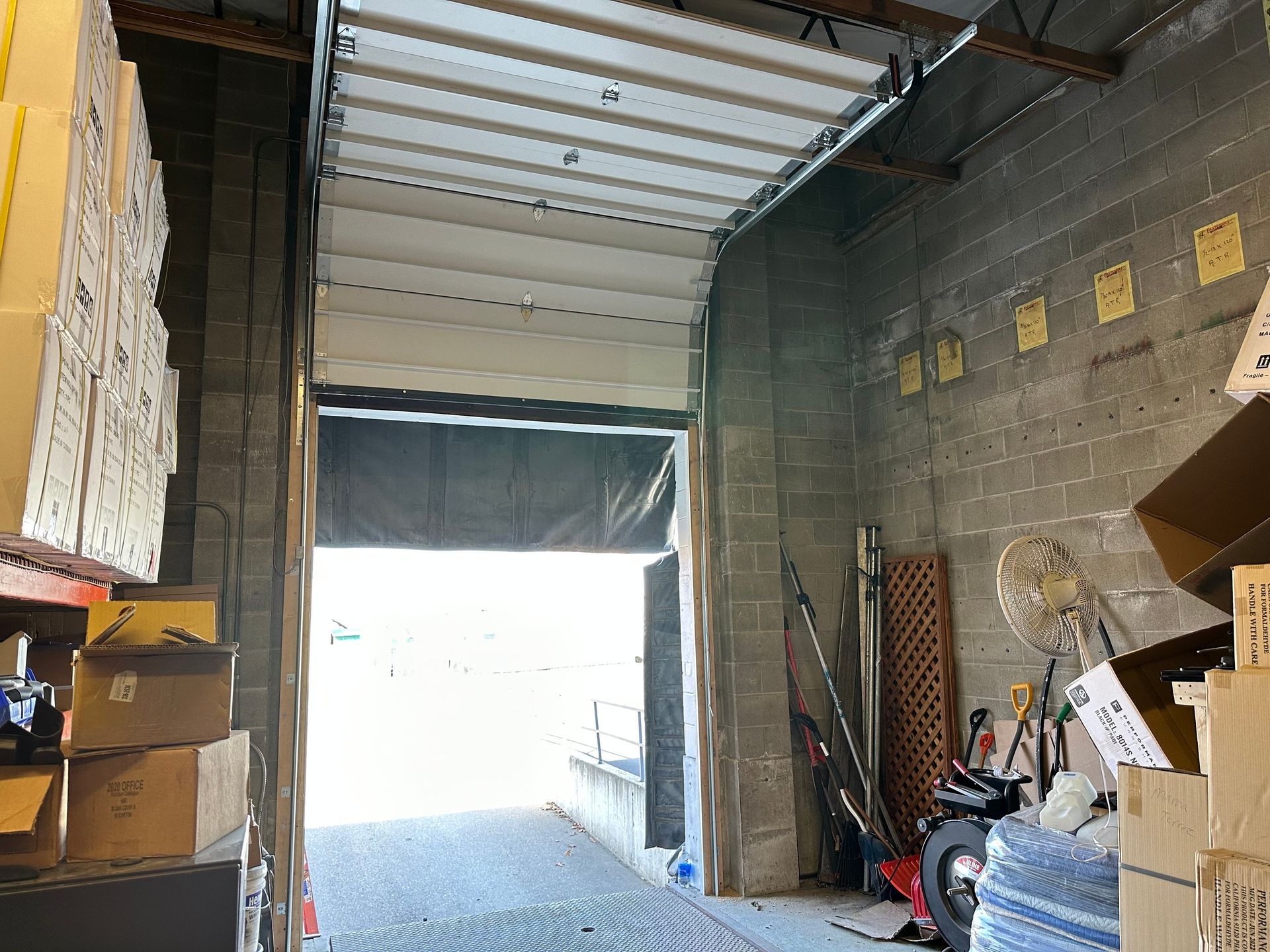 Inside a warehouse, a large roll-up door is partially open, revealing a bright outdoor area. Boxes stack on the left.