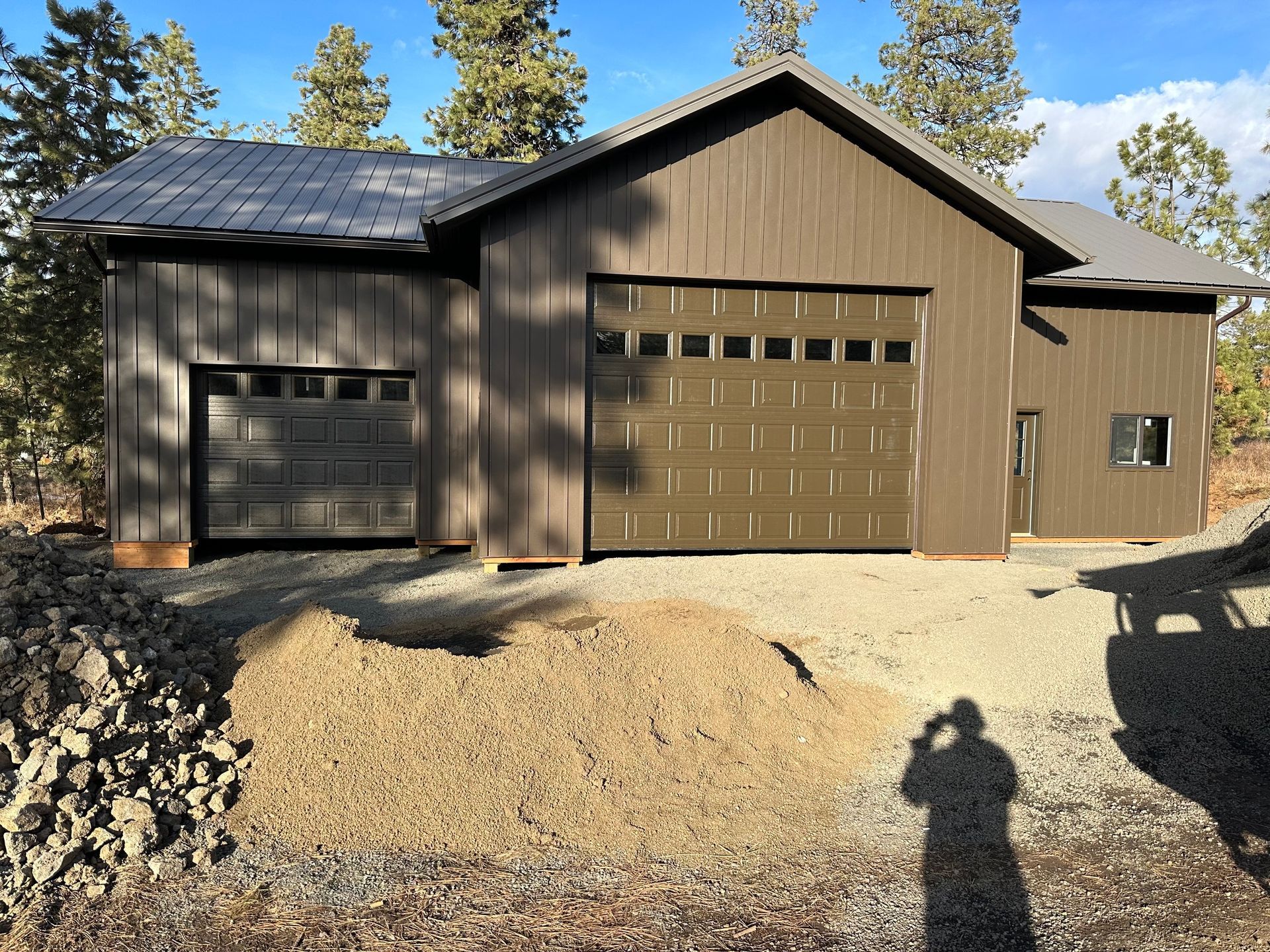 A brown, two-bay garage with vertical siding and metal roofing sits in a forest clearing next to a pile of gravel.