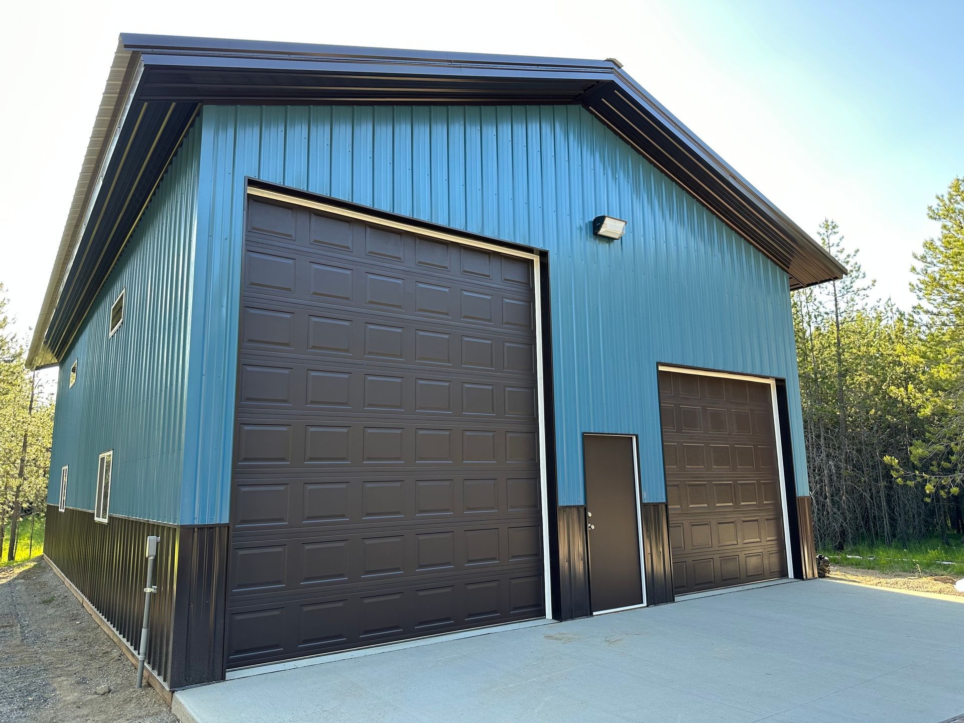 A blue metal garage with two dark brown garage doors, a matching entry door, and dark trim, set in a wooded area.