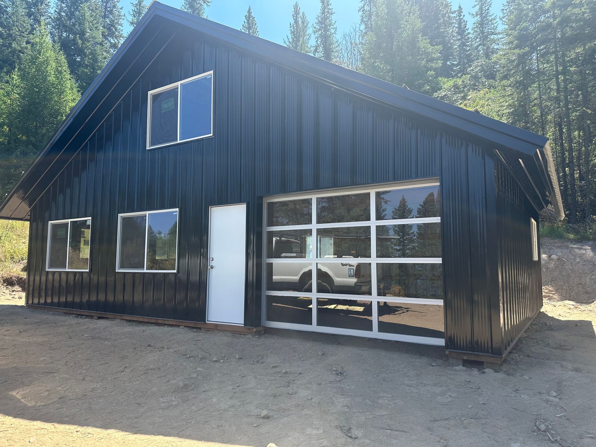 A black metal barn-style building with a glass garage door, a white entry door, and windows in a forest setting.
