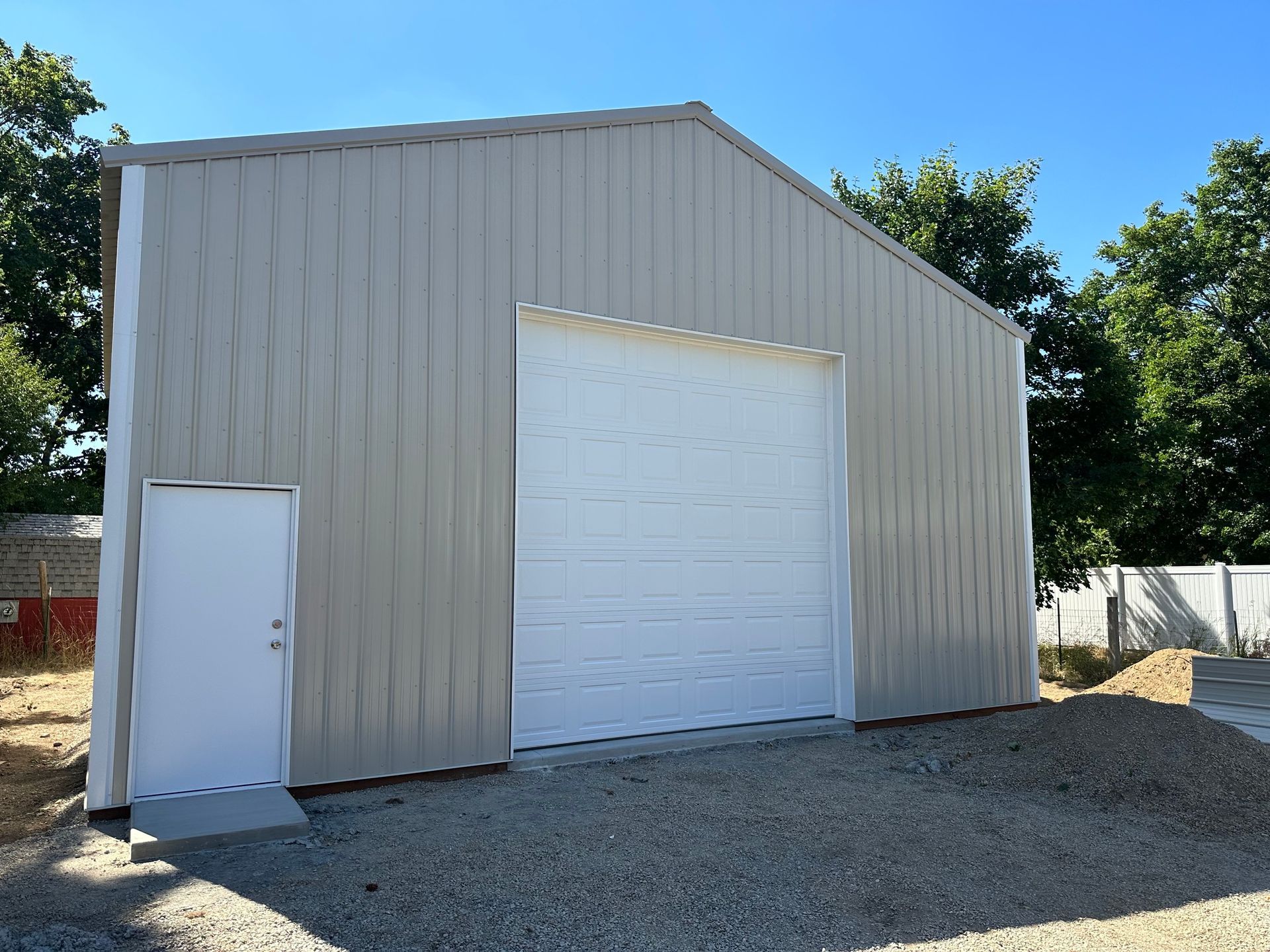 A tall, light-tan metal workshop with a white roll-up garage door and a side entry door on a gravel lot under a blue sky.