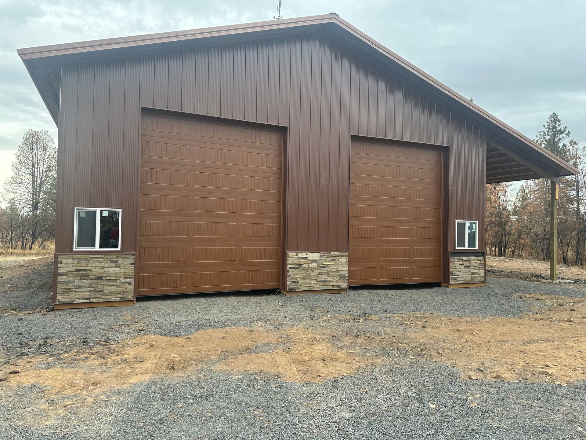 Brown metal barn with two garage doors, stone wainscoting, and windows on a gravel lot under a cloudy sky.