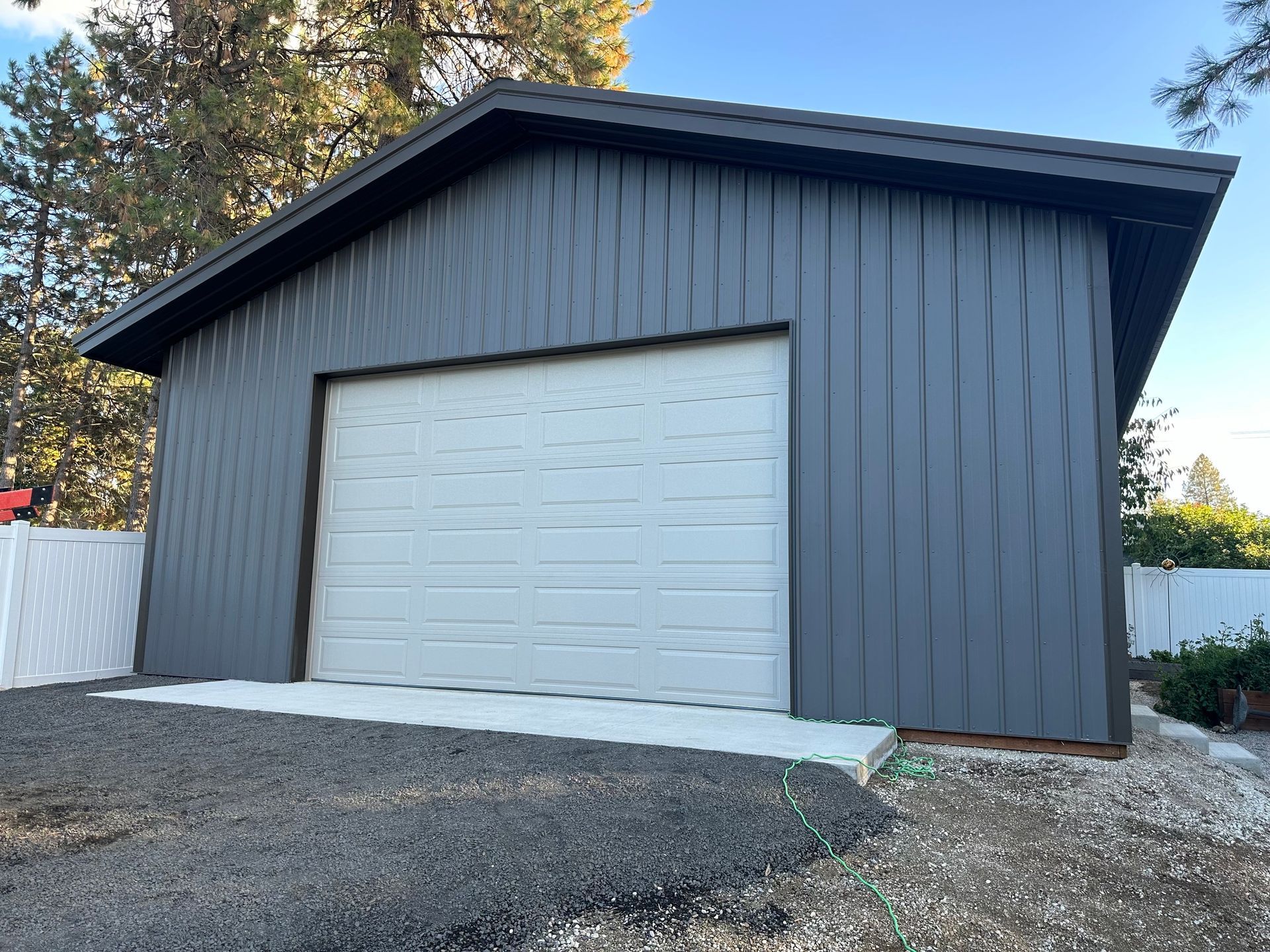 A detached dark gray garage with a white roll-up door, situated on a gravel lot near a white fence and pine trees.