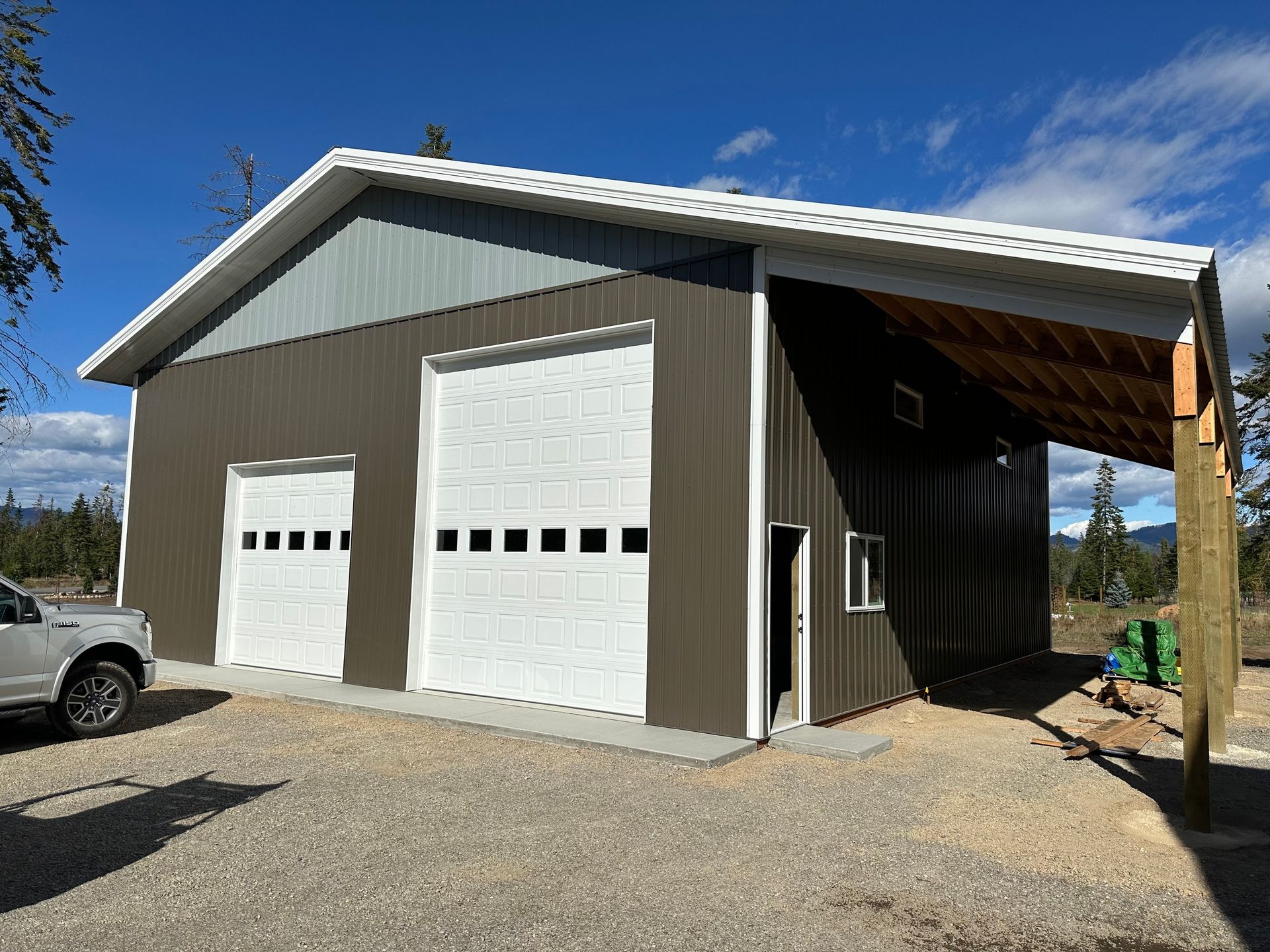 A brown metal pole barn with two white garage doors, a small side entry, and a large covered lean-to over gravel ground.