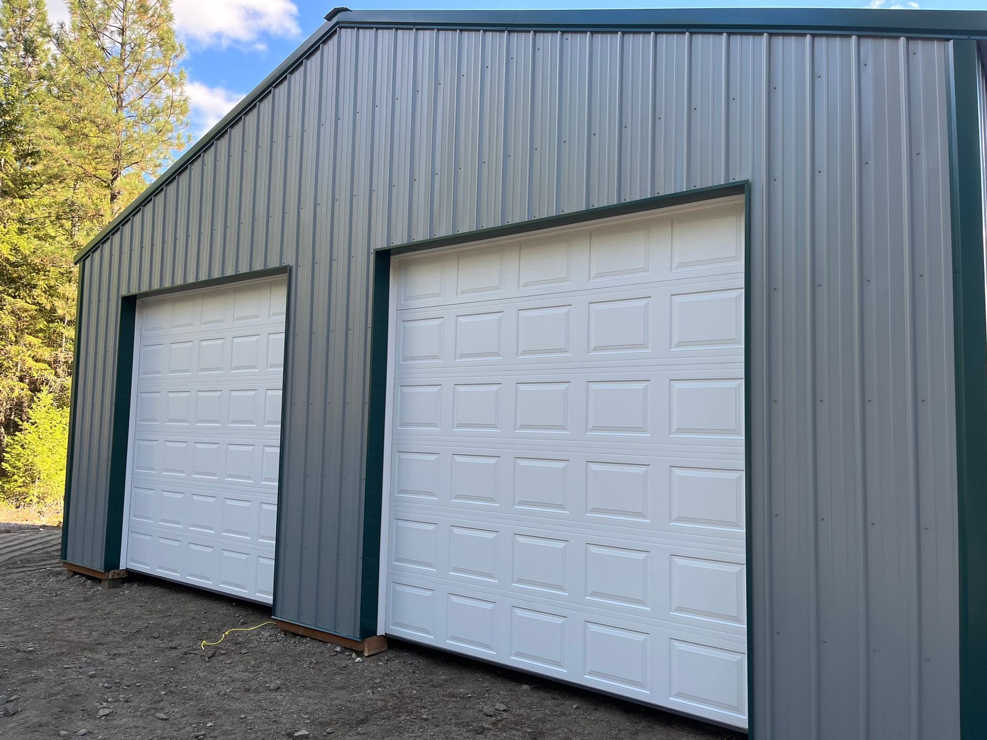 Two white garage doors set into the side of a grey metal-sided building near a forest.