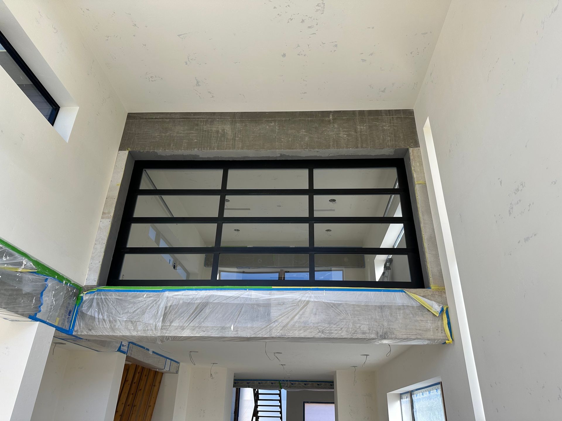 An interior view of a room under construction, featuring a large black metal-framed window partition on an upper level.