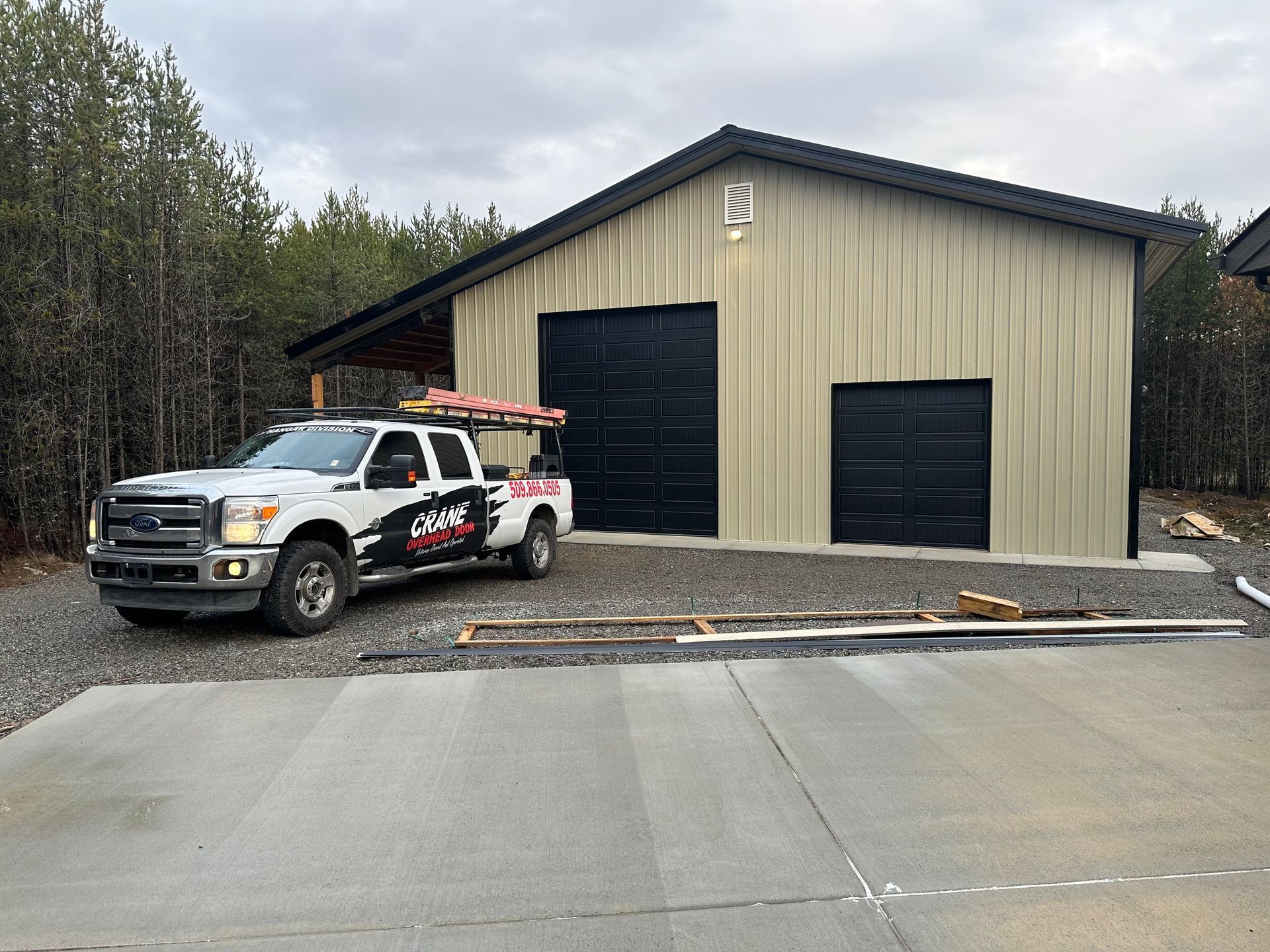 A white pickup truck with company decals parked on a gravel lot in front of a tan metal building with two black doors.