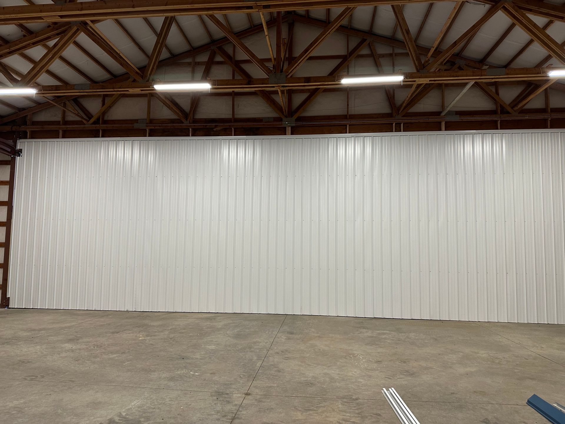 A white, vertically corrugated metal wall inside a large storage shed with exposed wooden roof trusses and overhead lights.