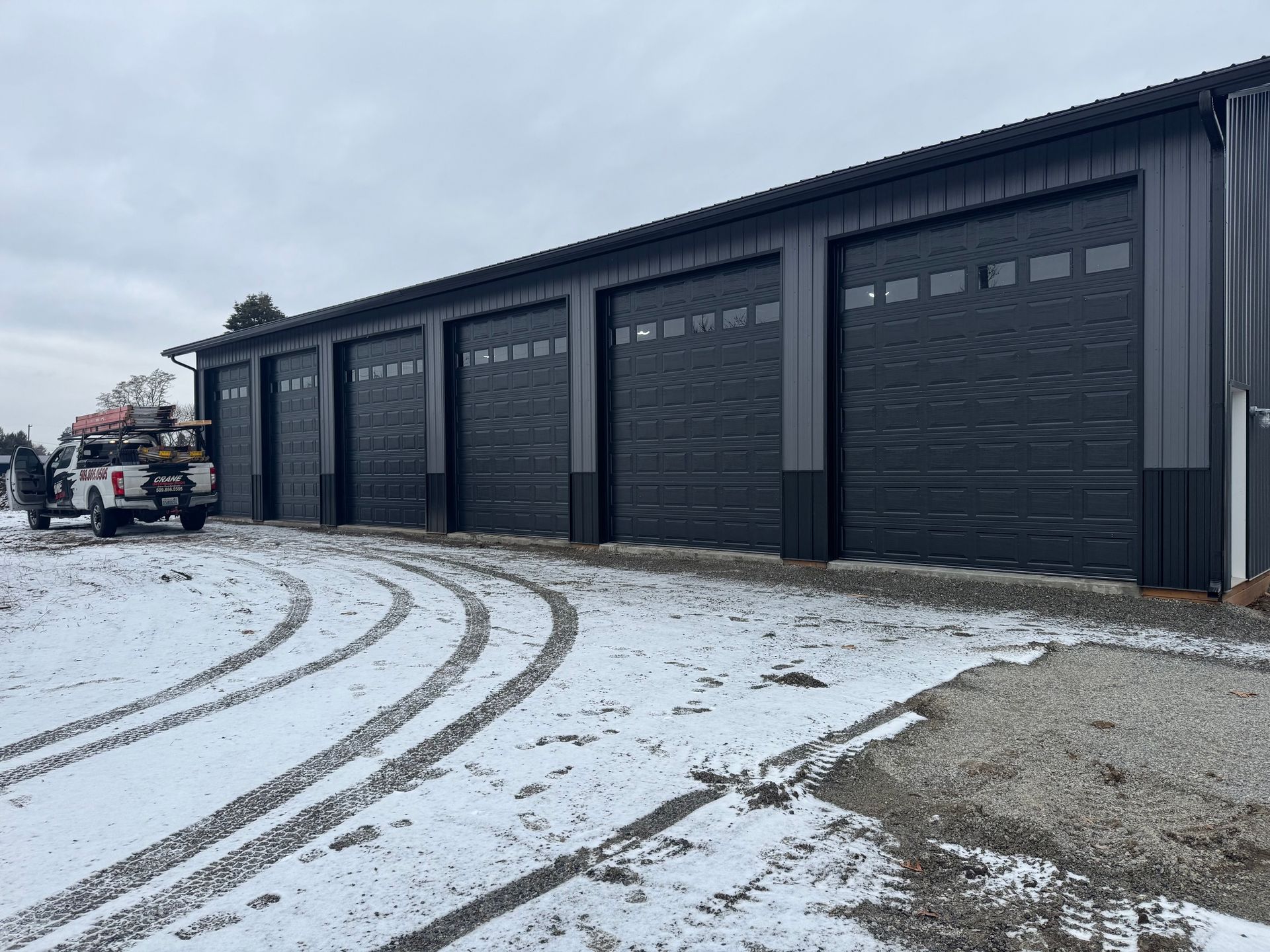 A row of six black garage doors on a grey metal building, with a work truck parked on a snowy gravel lot.