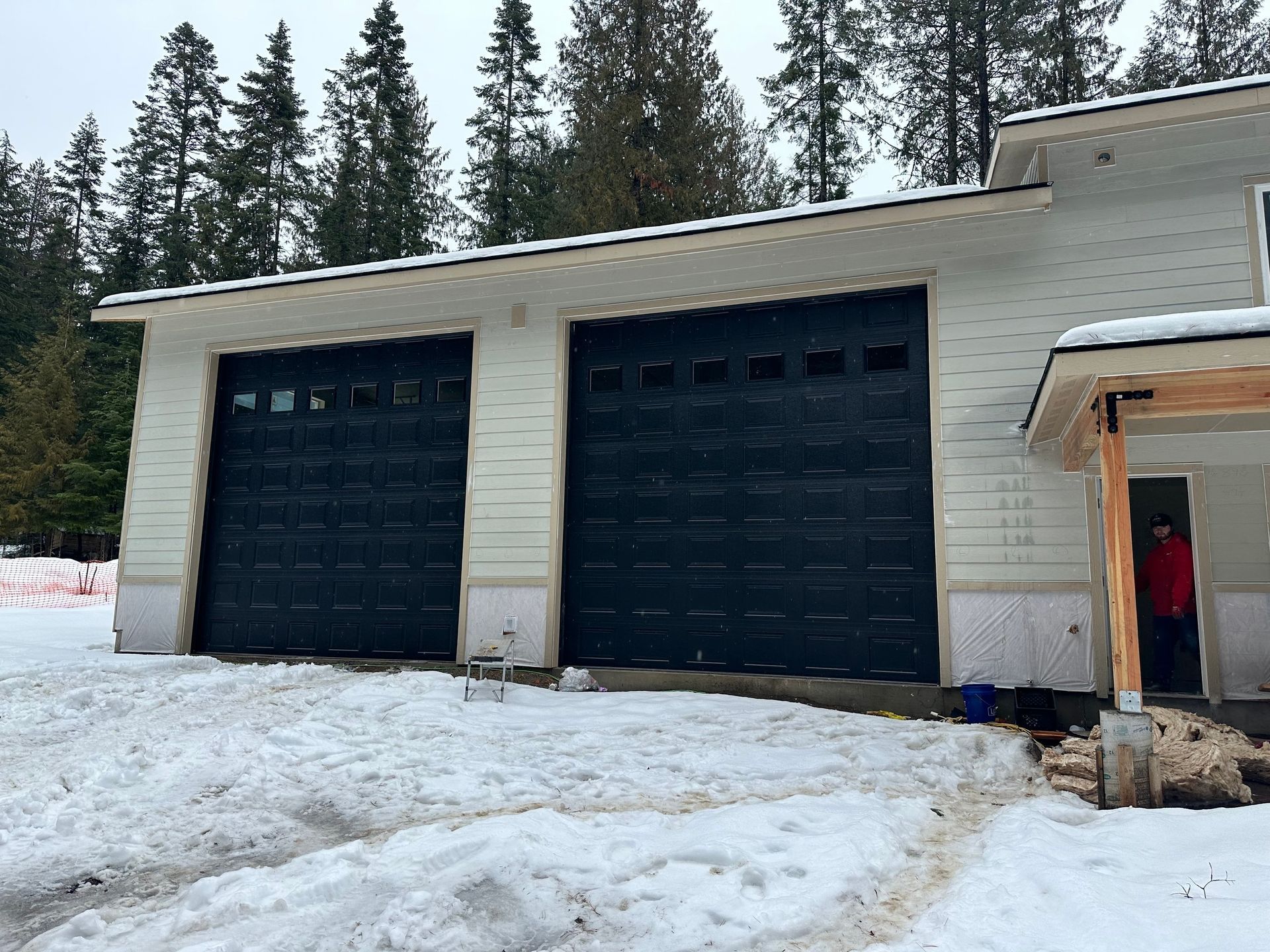 A light-colored house with two dark garage doors sits in a snowy, wooded yard with an unfinished entrance porch.