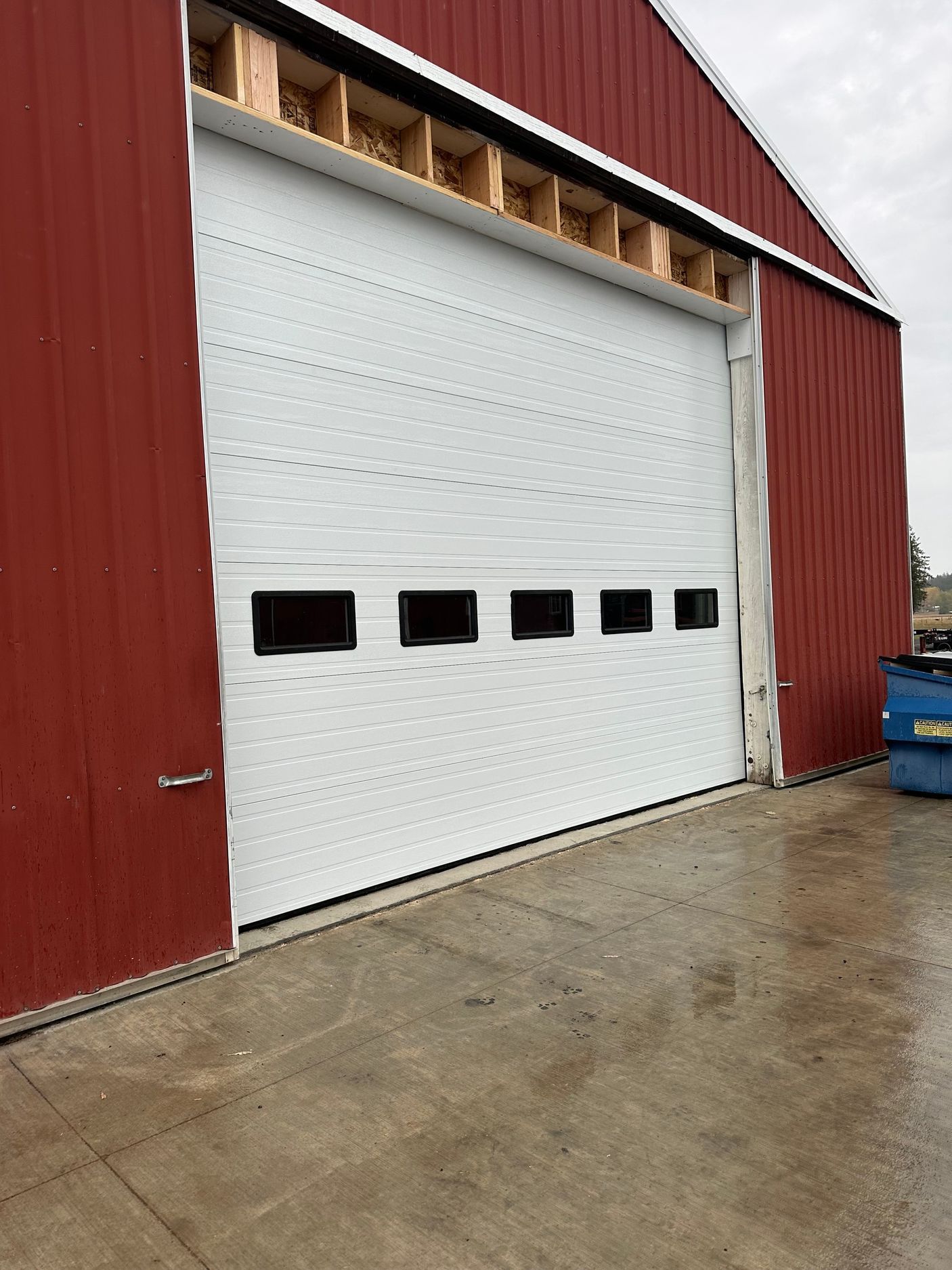 A white, multi-paneled garage door with a row of five windows, installed on a red metal agricultural building.