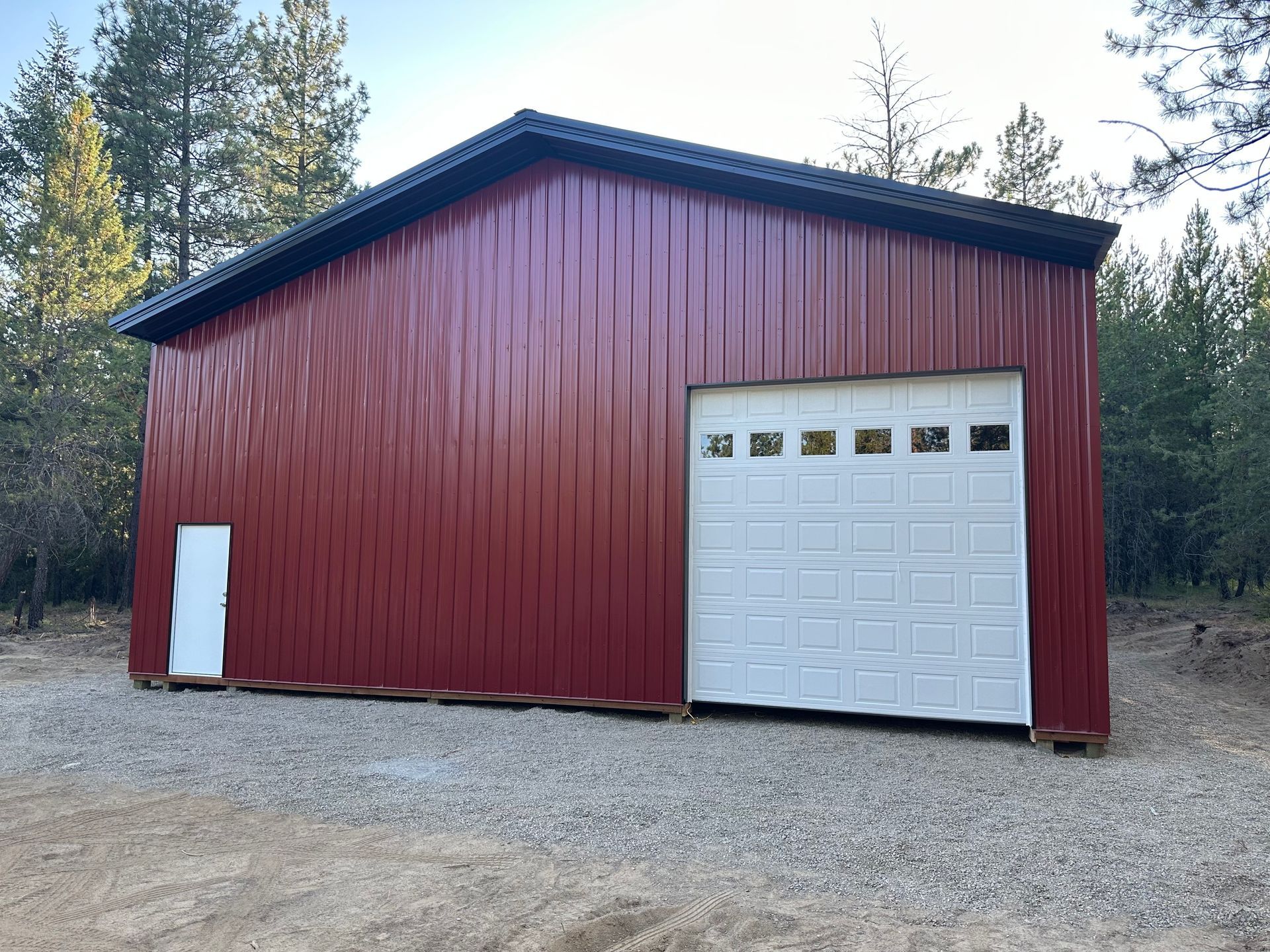 A red metal pole barn with a white garage door and a side entry door, situated in a wooded area on a gravel lot.