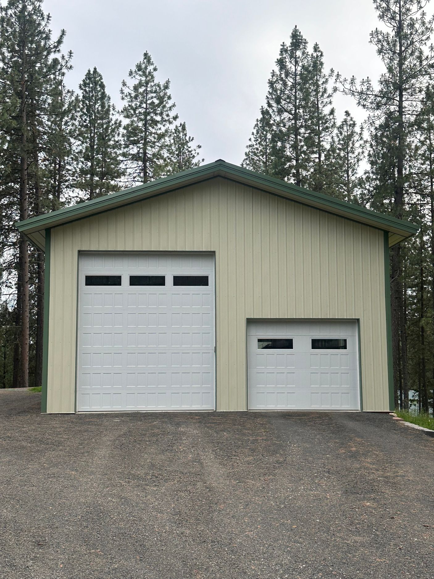 A detached garage with light beige siding and green trim, featuring one large and one small white garage door in a forest.