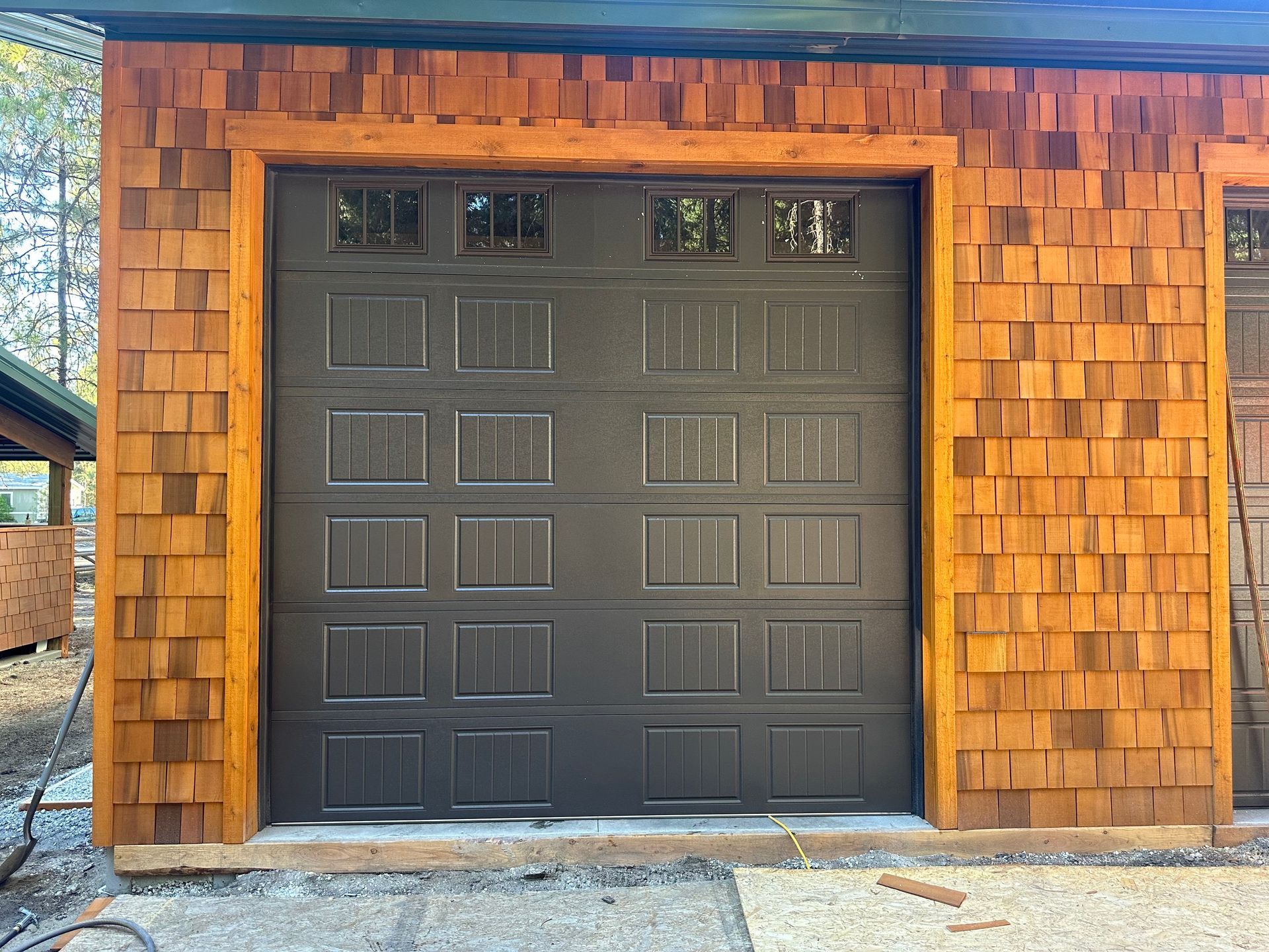 A black garage door with four top-row windows, framed by light-colored cedar shake siding on a building exterior.
