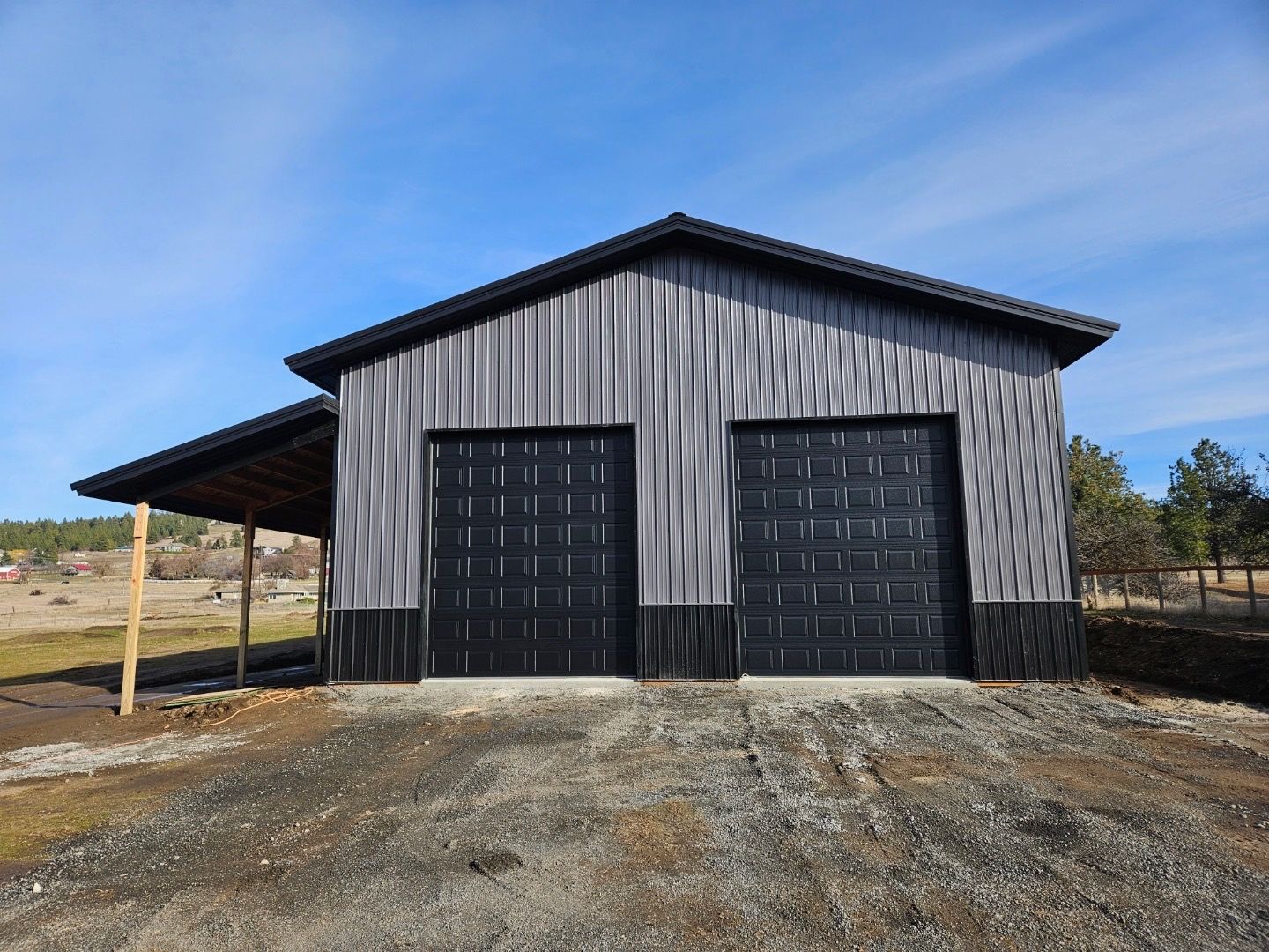 A gray, metal-sided barn with two black garage doors and a side lean-to, situated on a gravel lot under a blue sky.