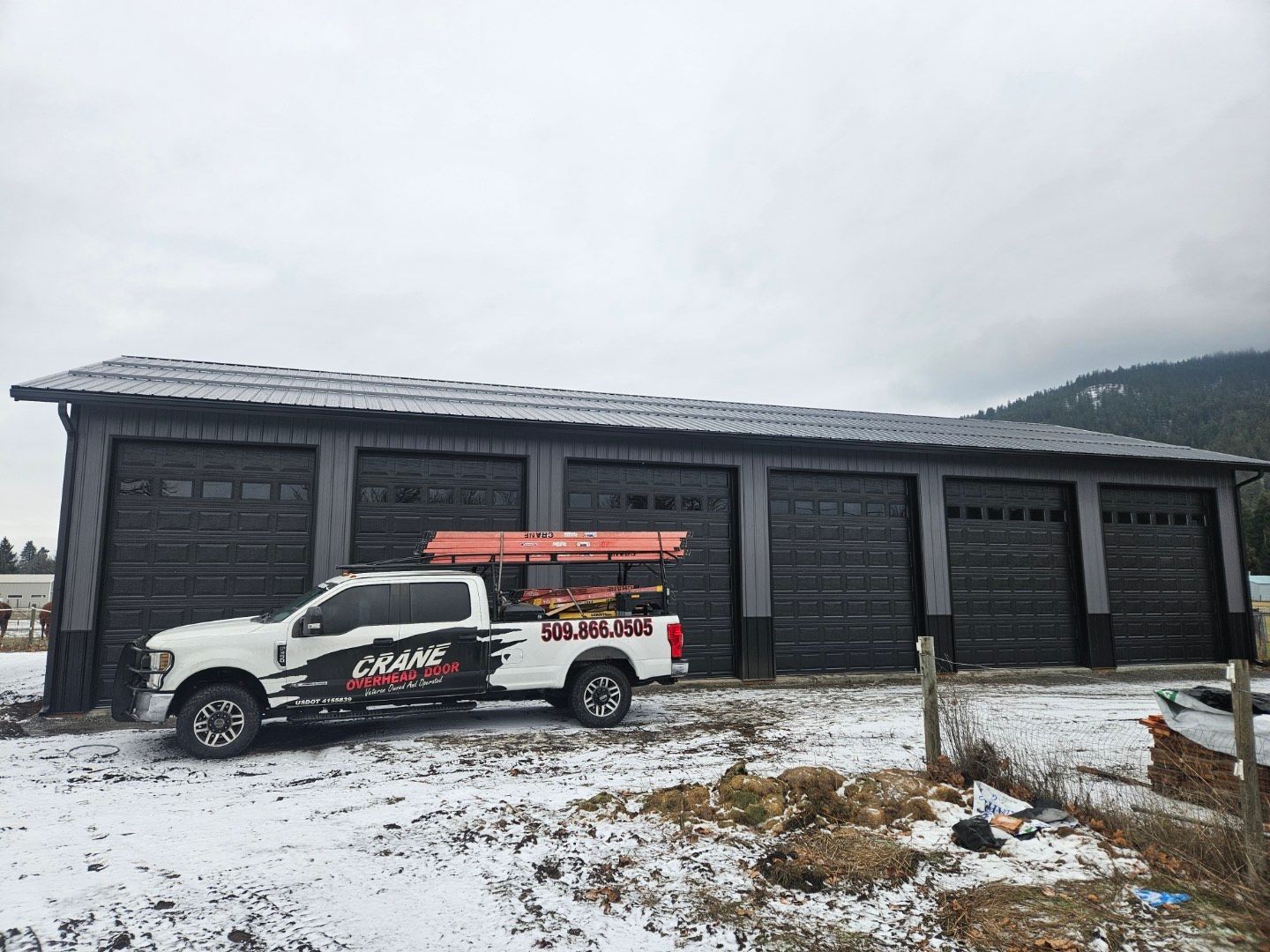 A white service truck with an orange ladder rack parked in front of a long garage building with five black roll-up doors.