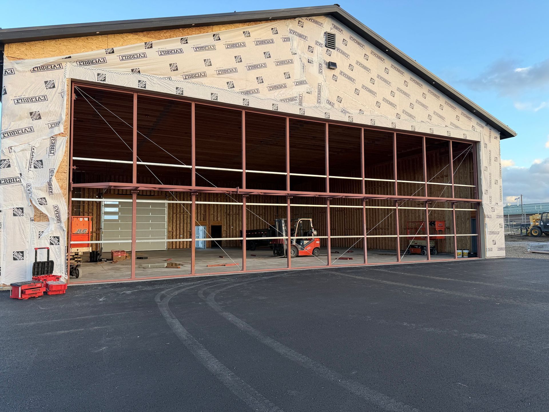 A large building under construction featuring an expansive open doorway with a steel frame, viewed from an asphalt lot.