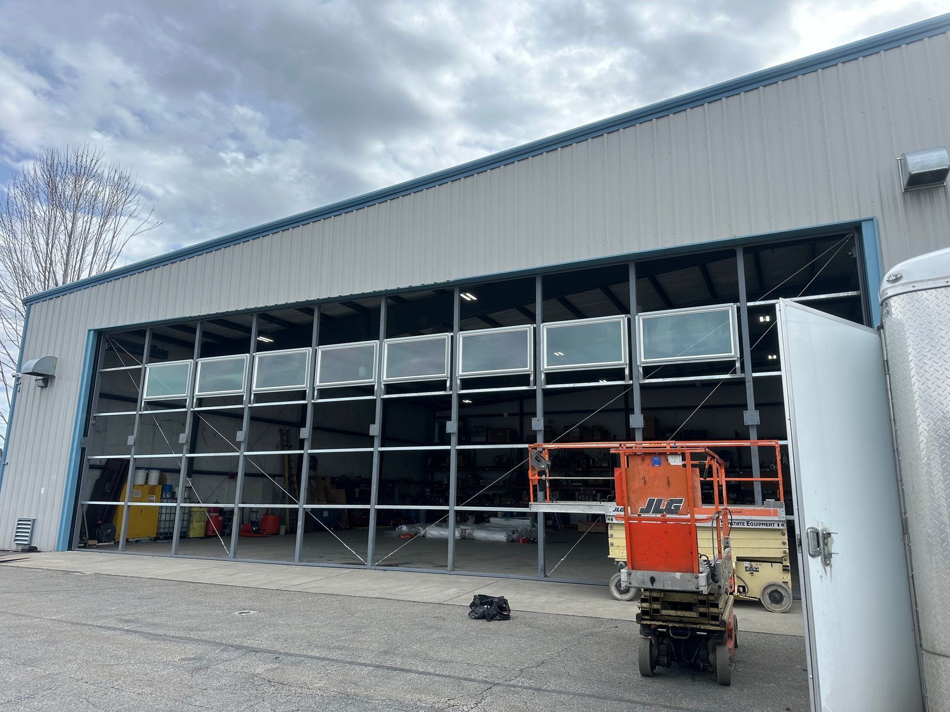 A large warehouse building with an open bay door and a yellow scissor lift parked in front under a cloudy sky.
