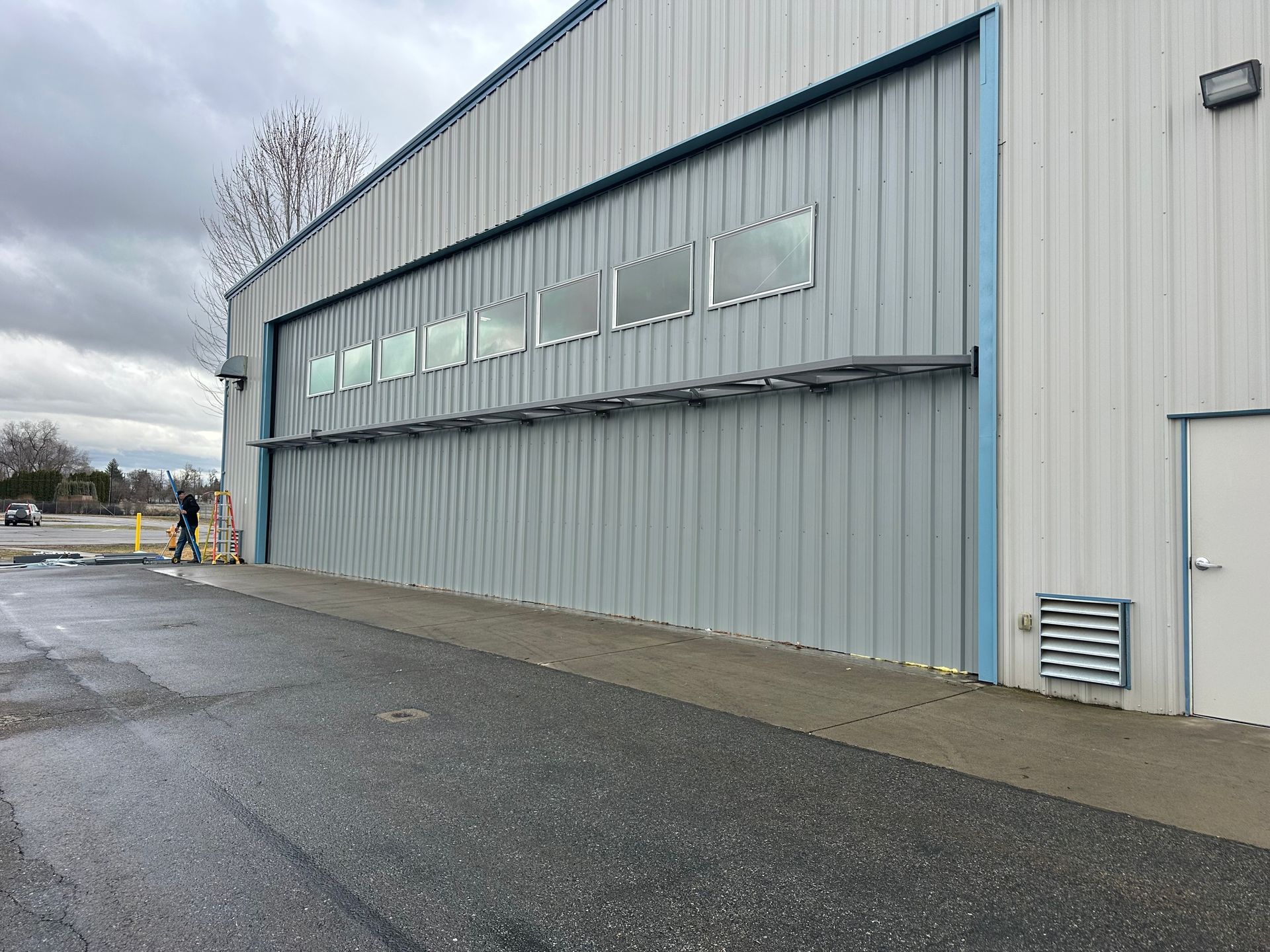 A large, light-gray industrial metal building with a wide horizontal hangar door featuring a row of windows.