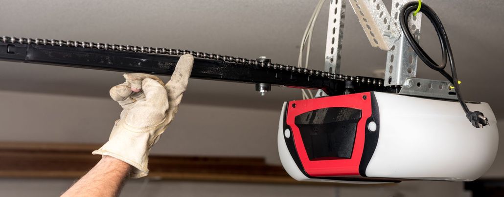 A gloved hand points to the chain and rail of a white and red garage door opener mounted to a ceiling.