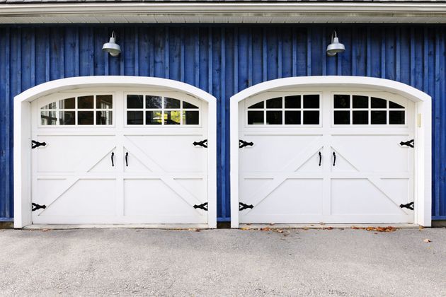 Two white arched garage doors with black hinges and windows set against a vertical blue wood-paneled wall.