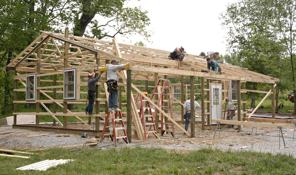 Construction workers build the wooden frame of a house on a rural property surrounded by trees.