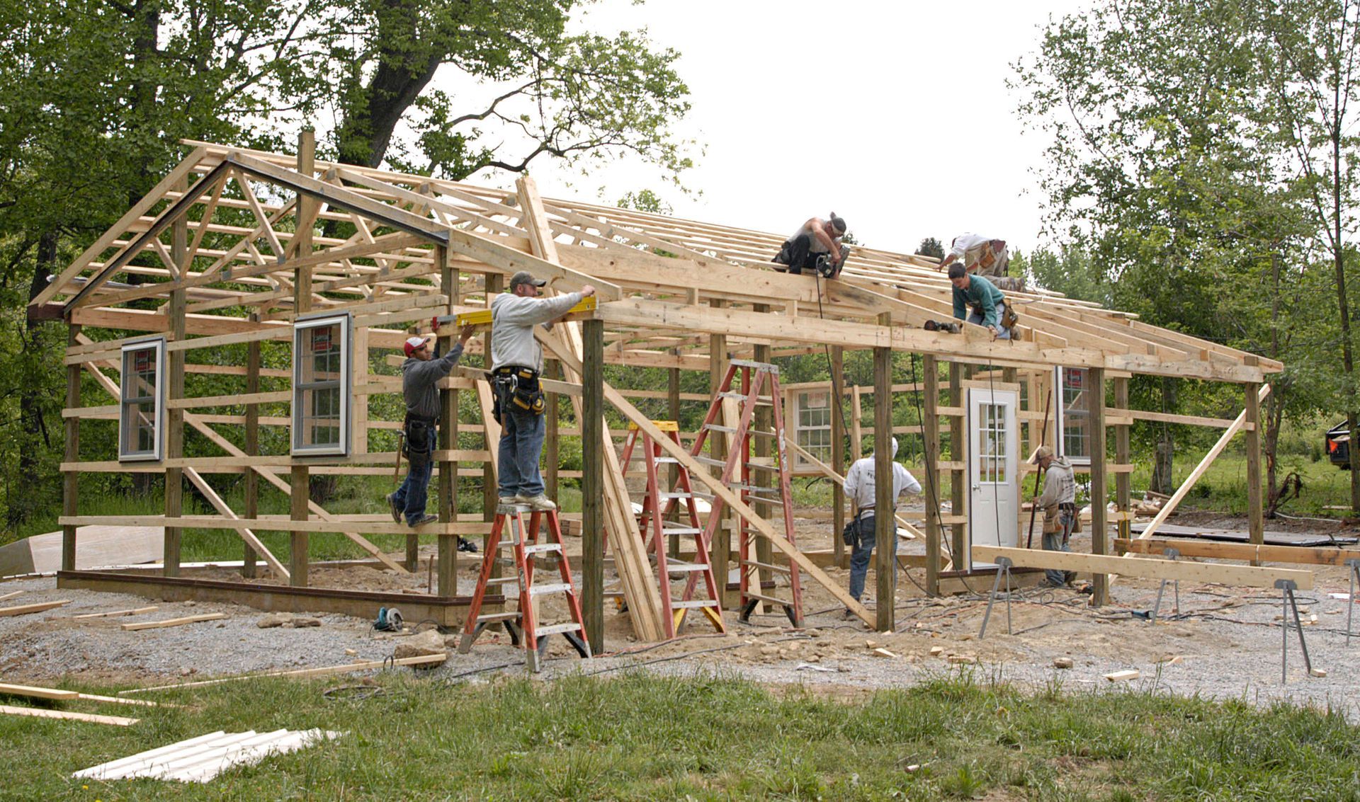 Construction workers build the wooden frame of a house on a rural property surrounded by trees.