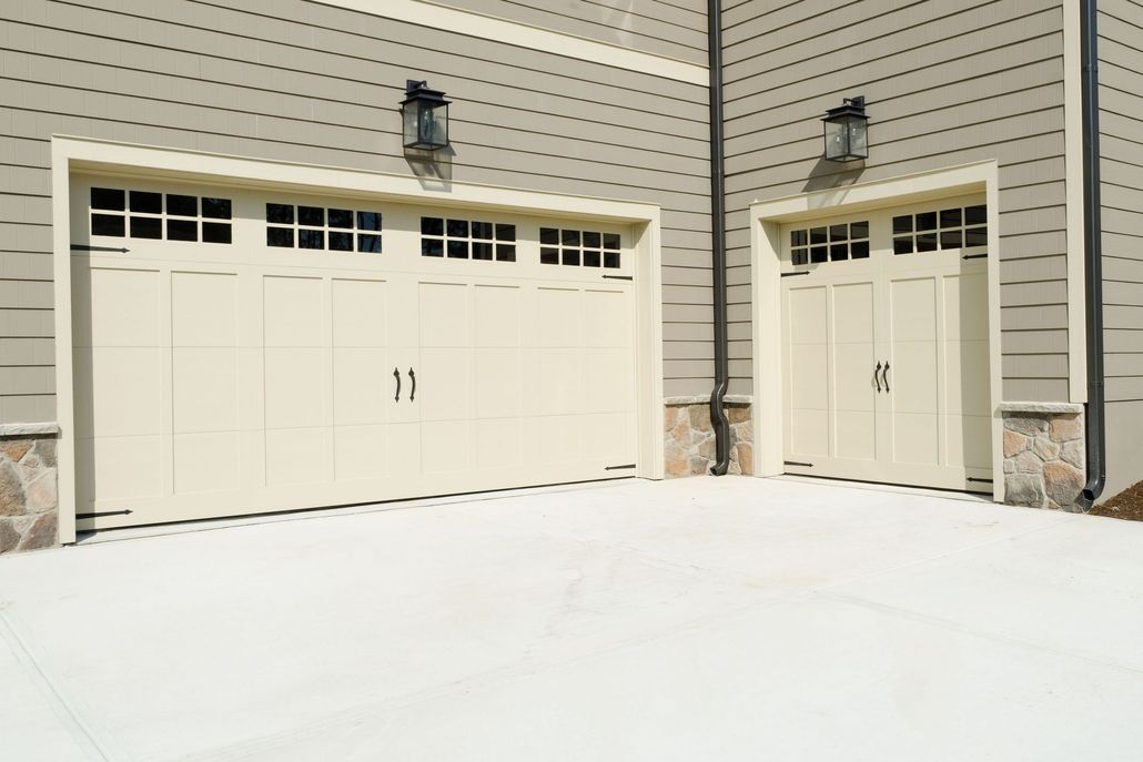 Cream-colored double and single garage doors with window panels installed on a house with beige siding.