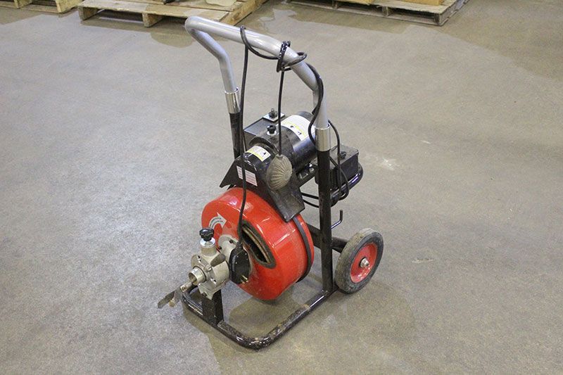 Red and black electric drain cleaner on wheels. Gray handle, industrial setting.