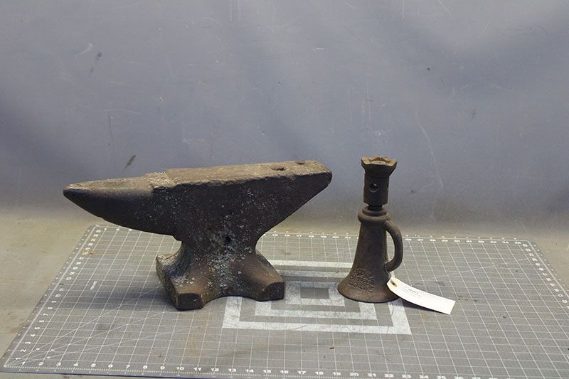 An anvil and a small candlestick on a gridded surface. Both are dark and metallic.