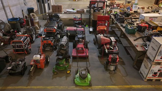 A cluttered workshop displays various lawnmowers, tractors, and equipment on a concrete floor.