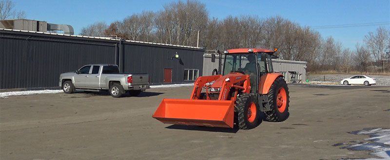 An orange tractor with a front loader on asphalt, with a silver truck and white car.