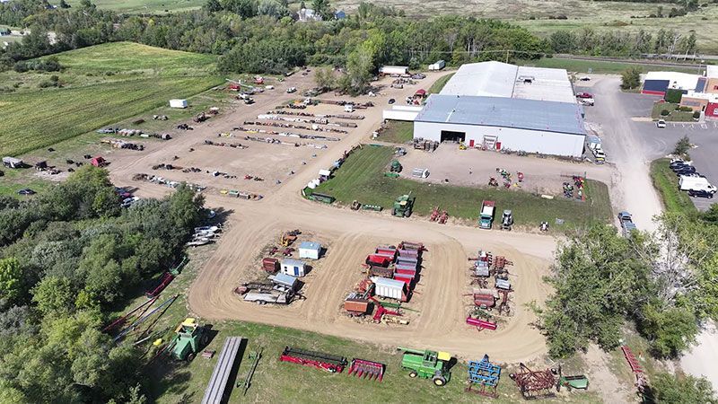 Overhead view of an agricultural equipment auction. Tractors, implements, and trailers are displayed outdoors near a large building.