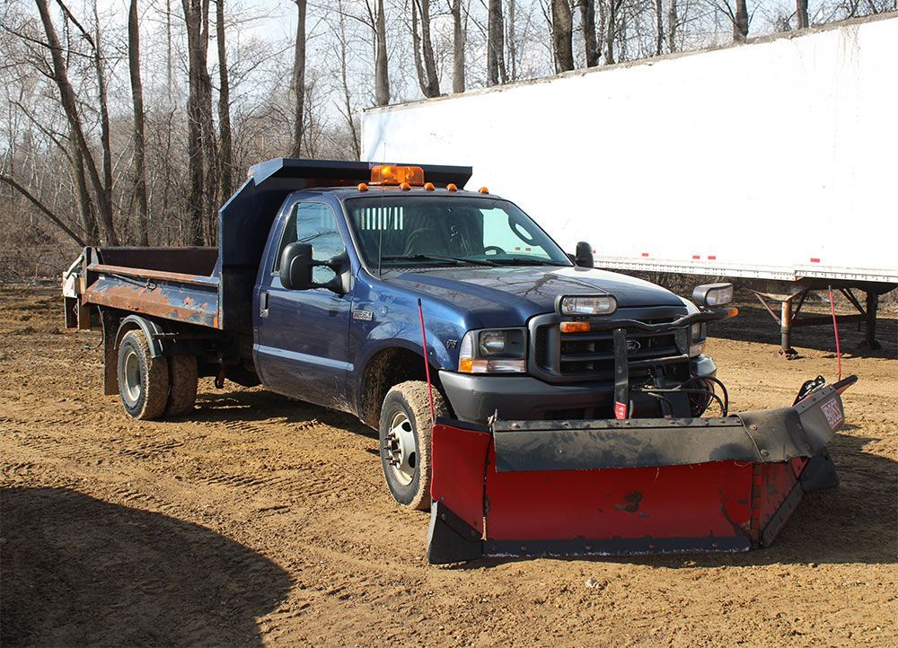 Blue pickup truck with snowplow, dump bed, and flashing lights, parked on dirt.