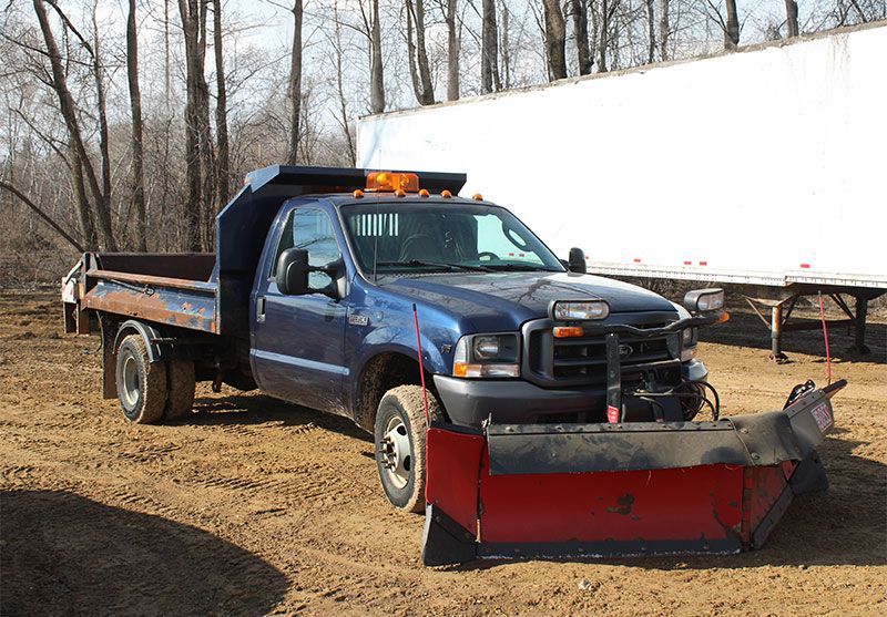 Blue pickup truck with a snowplow on a dirt lot.