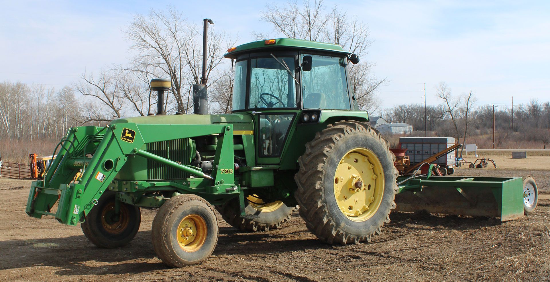 Green John Deere tractor with a front loader and a box blade, on a dirt field.