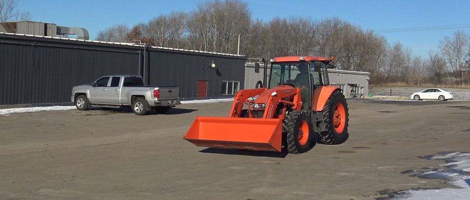 Orange tractor with a front-end loader on a paved lot with a silver truck and a white car.