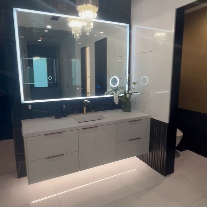 Modern bathroom vanity with backlit mirror, white cabinets, black wall accents, and chandelier lighting