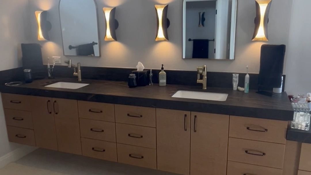 Modern bathroom vanity with double sinks, mirrors, wall sconces, and beige cabinets under dark countertops.
