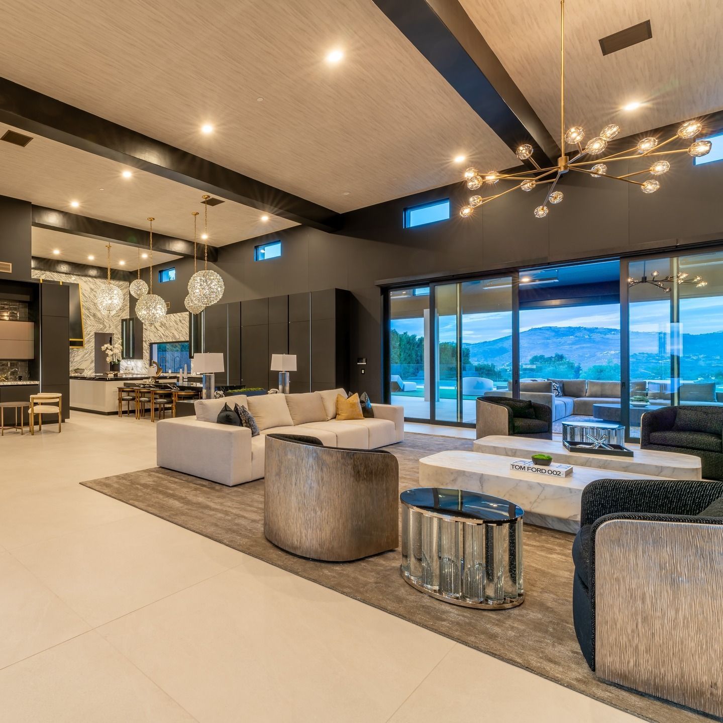 Modern luxury living room with beige seating, dark walls, pendant lights, and large glass doors overlooking a pool.