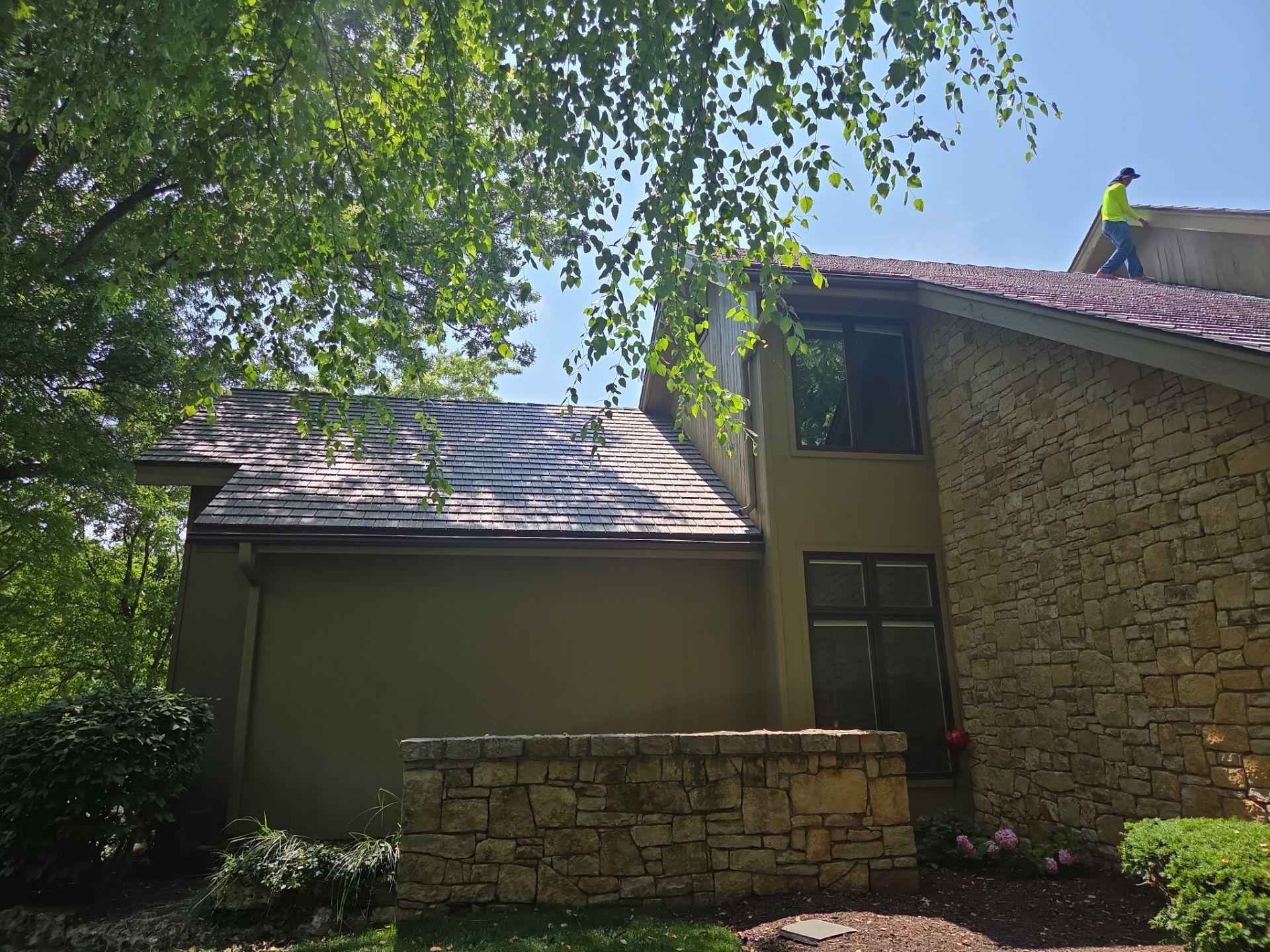 Person on roof of two-story house with a tan facade and stone accents on a sunny day.
