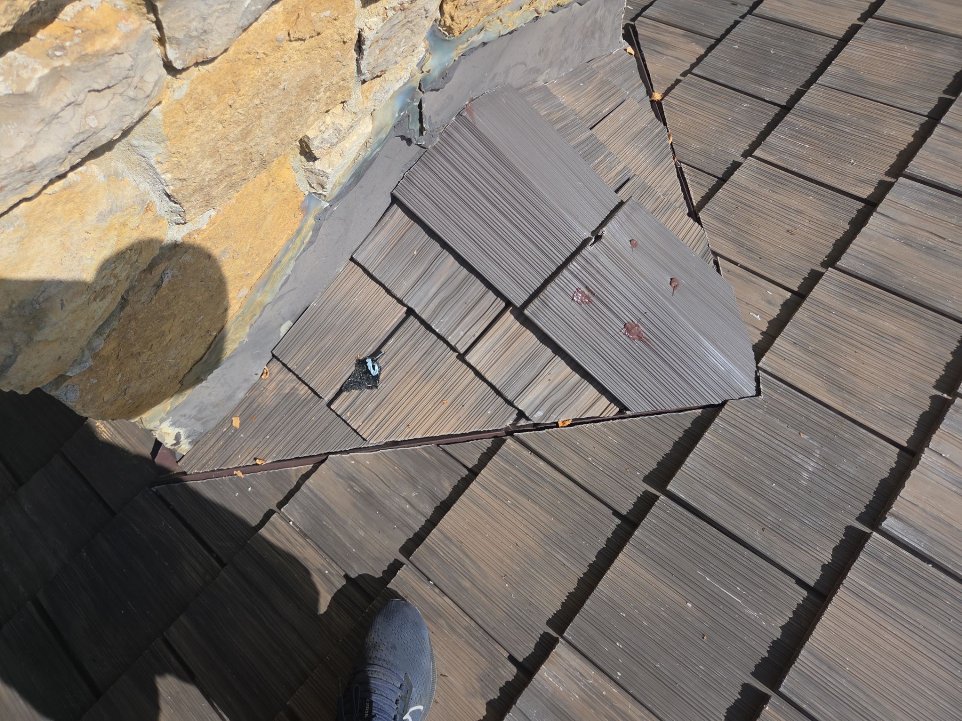 Close-up of damaged wood shingle roof near a stone wall. A foot is also visible.