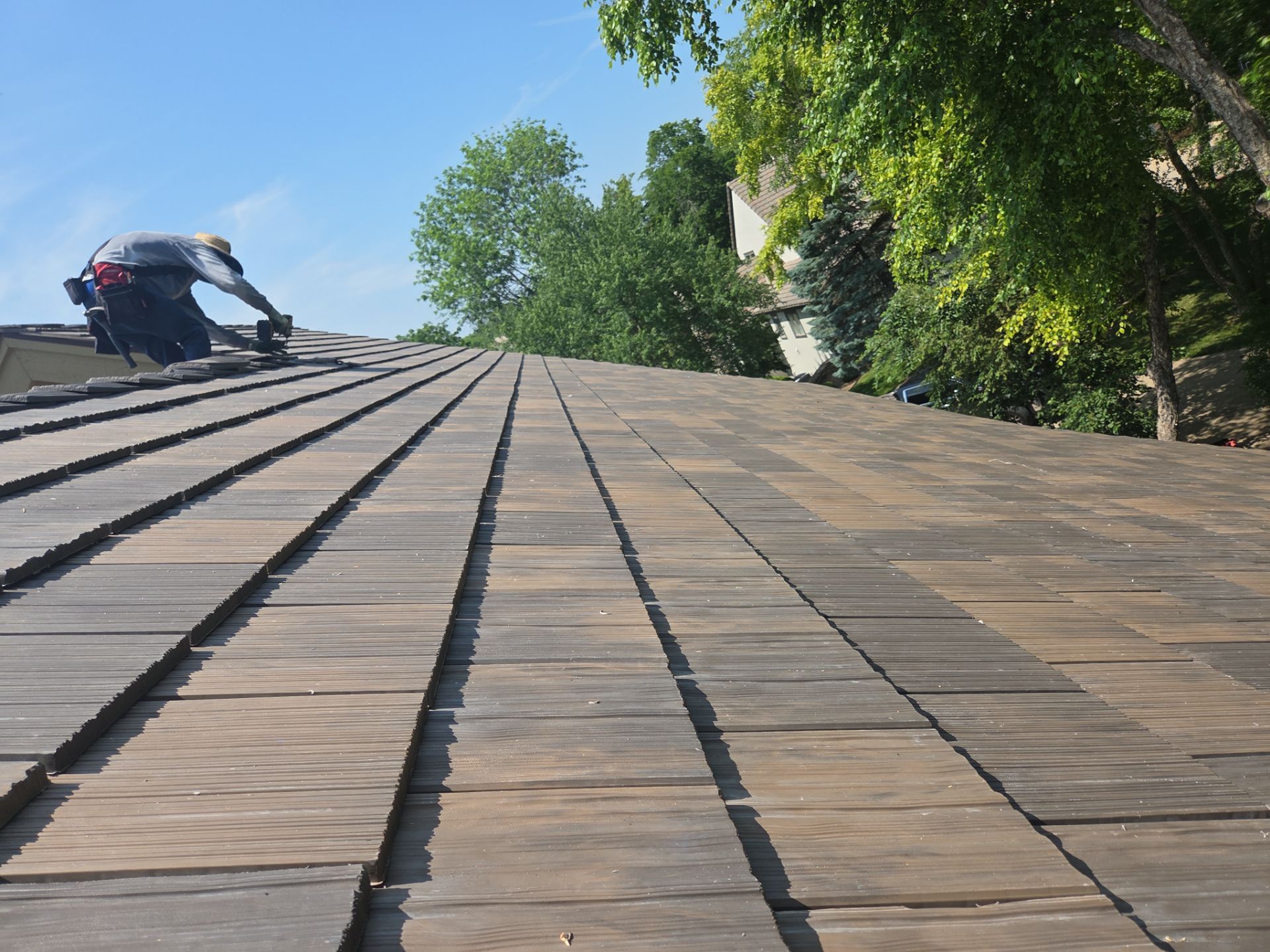 Roofer working on a wood shingle roof under a sunny blue sky, surrounded by green trees.