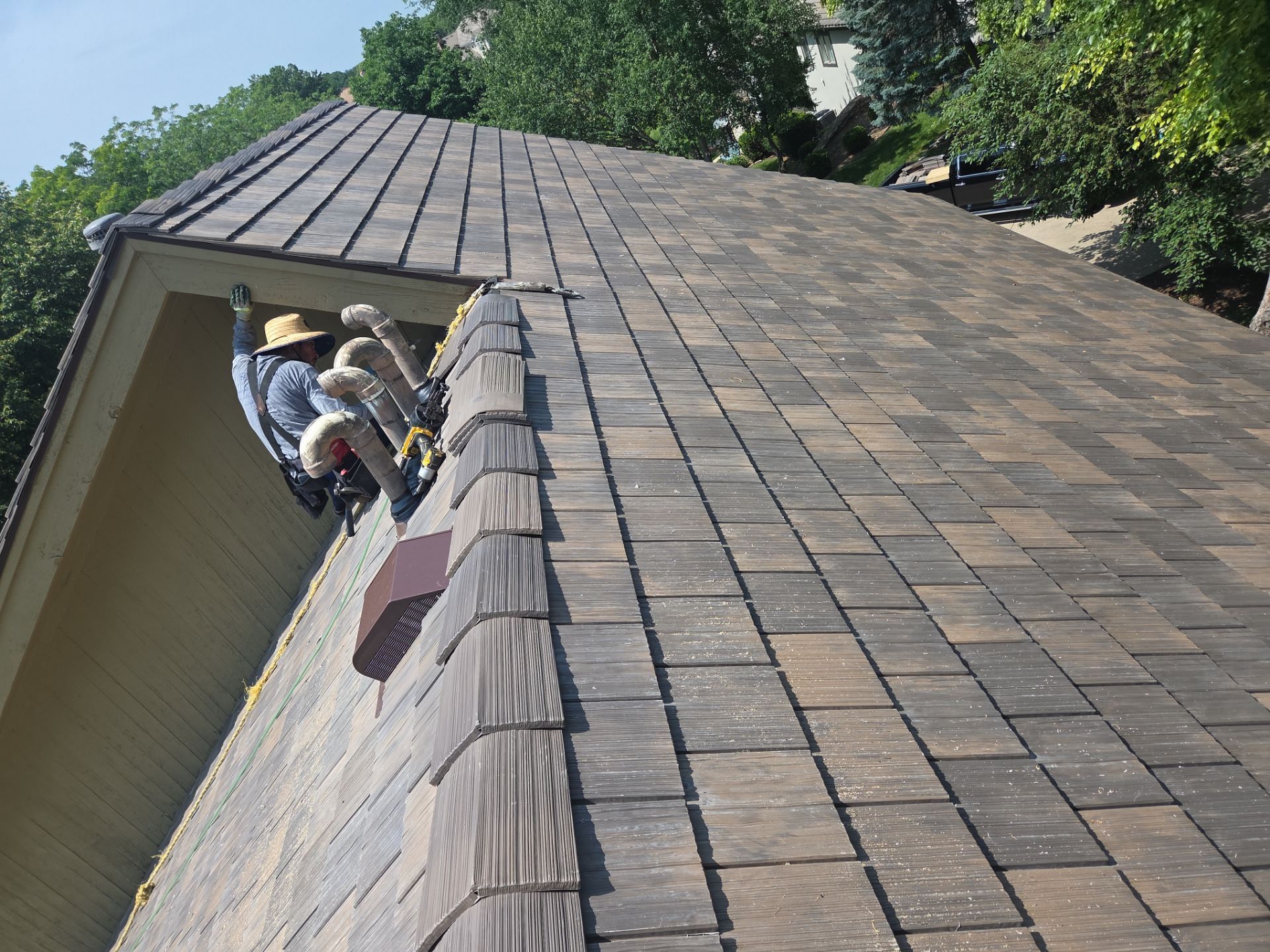 Workers on a roof installing brown shingles, trees in the background, sunny day.