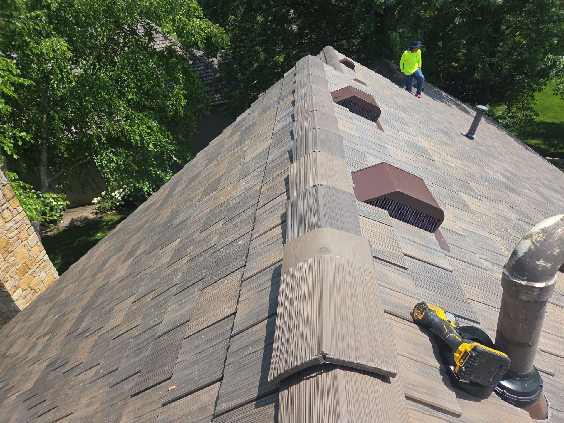 Roofer working on a multi-tone shingle roof; a yellow-clad person stands, and tools are visible.