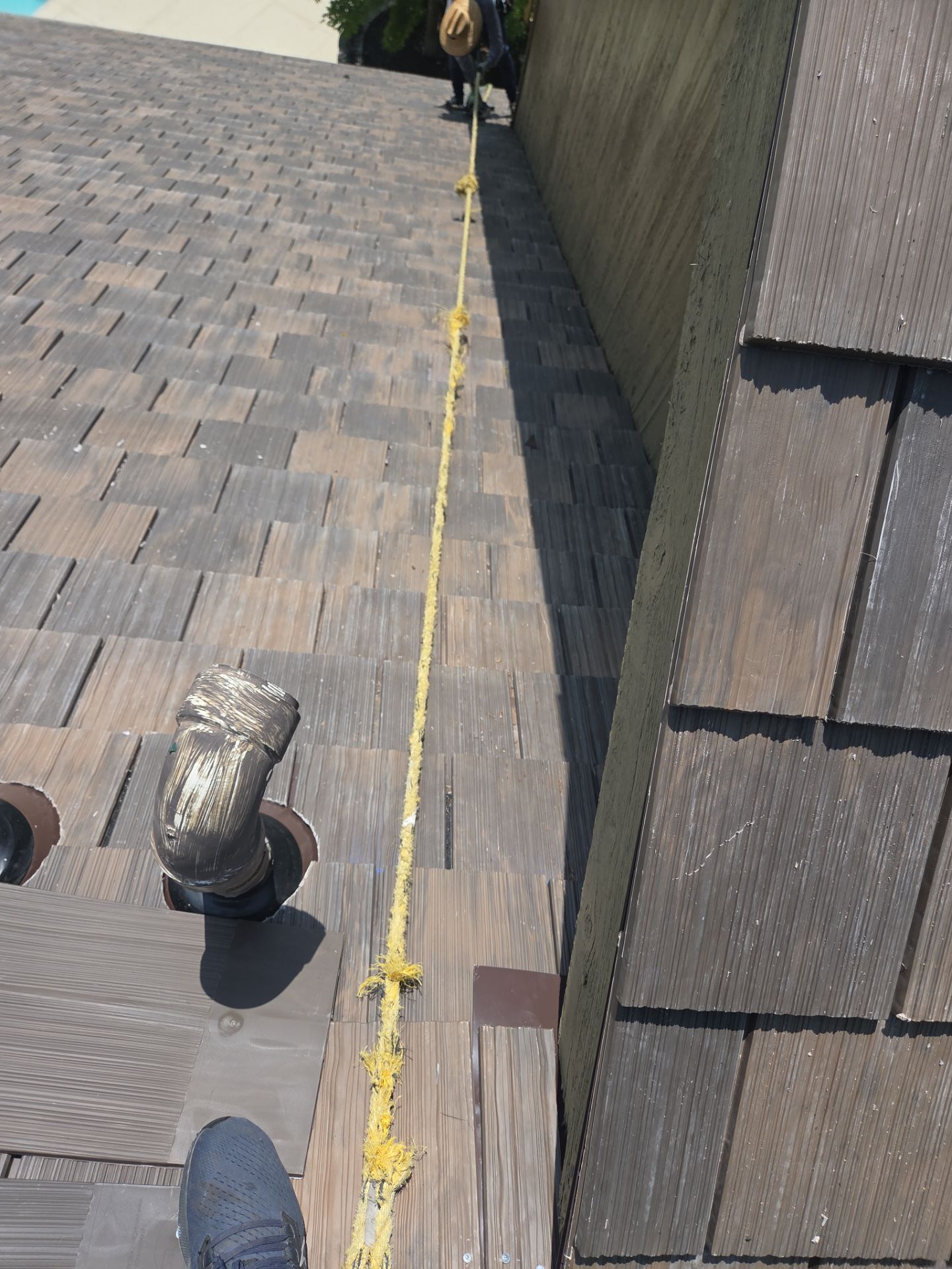 Person on a shingle roof, attached to a yellow safety line. A metal pipe is also visible.